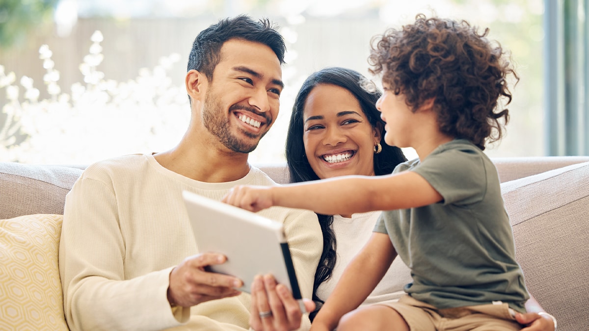 A young boy with his parents on the couch pointing at a tablet