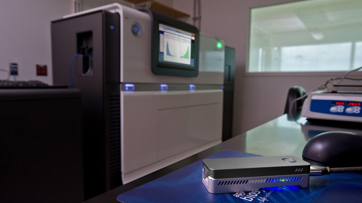 A small nanopore sequencer (about the size of a short stapler) sits on a table in the foreground. In the background is a large sanger sequencer machine (the size of a large copy machine)