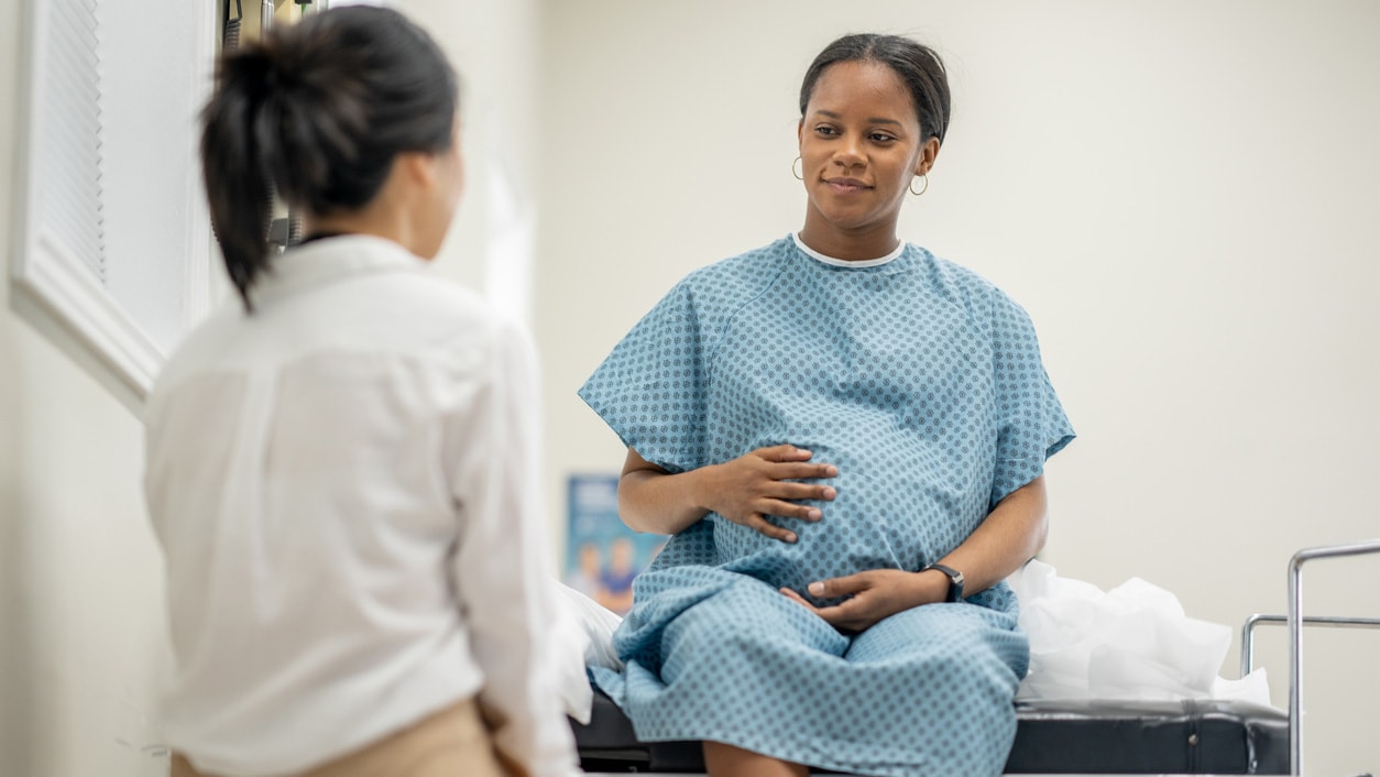 A pregnant woman, wearing a medical gown, sits on an exam table at a prenatal check-up. A doctor sits across from her.