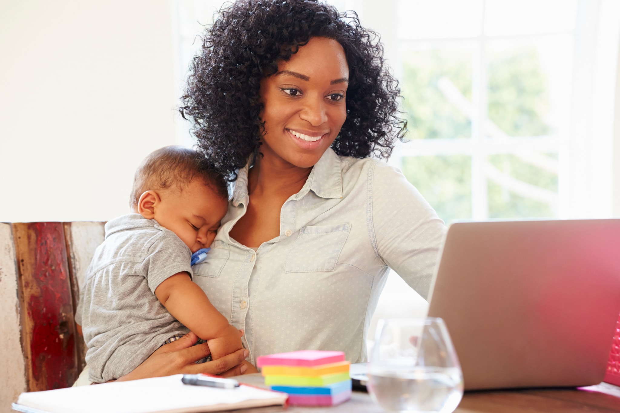 Woman holding sleeping baby while she uses her laptop.