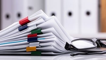 Stack of papers on a desk next to a pair of glasses.