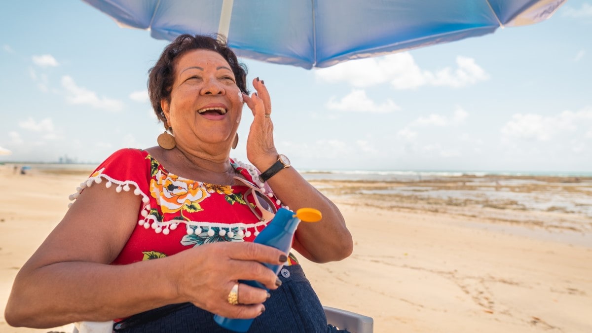 Photo of a woman sitting under an umbrella on the beach applying sunscreen