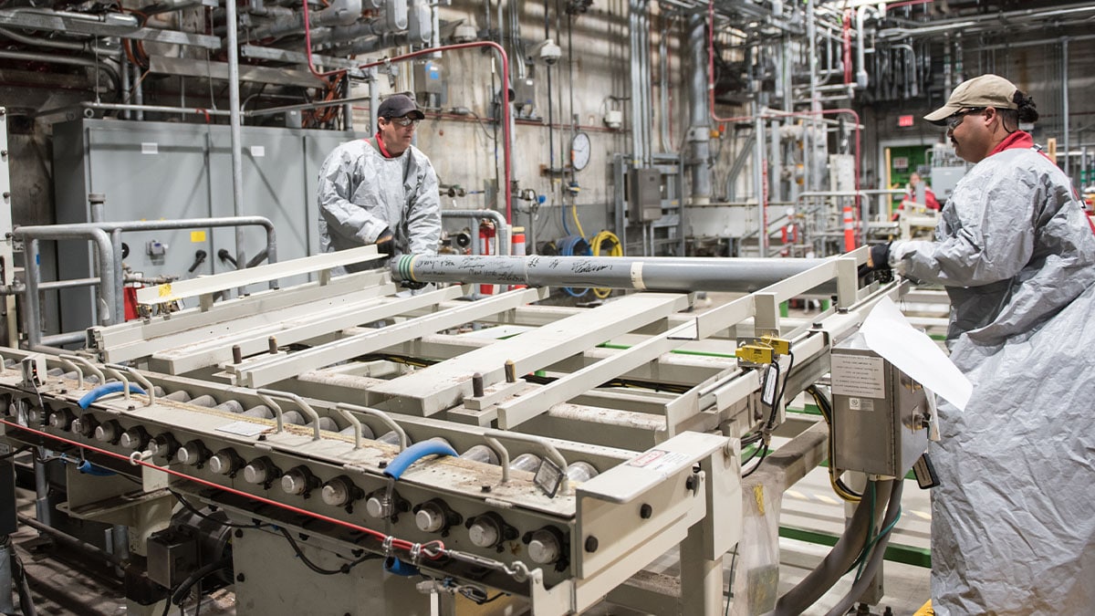 An image of two worker in personal protective equipment loading a chemical munition onto a conveyor.