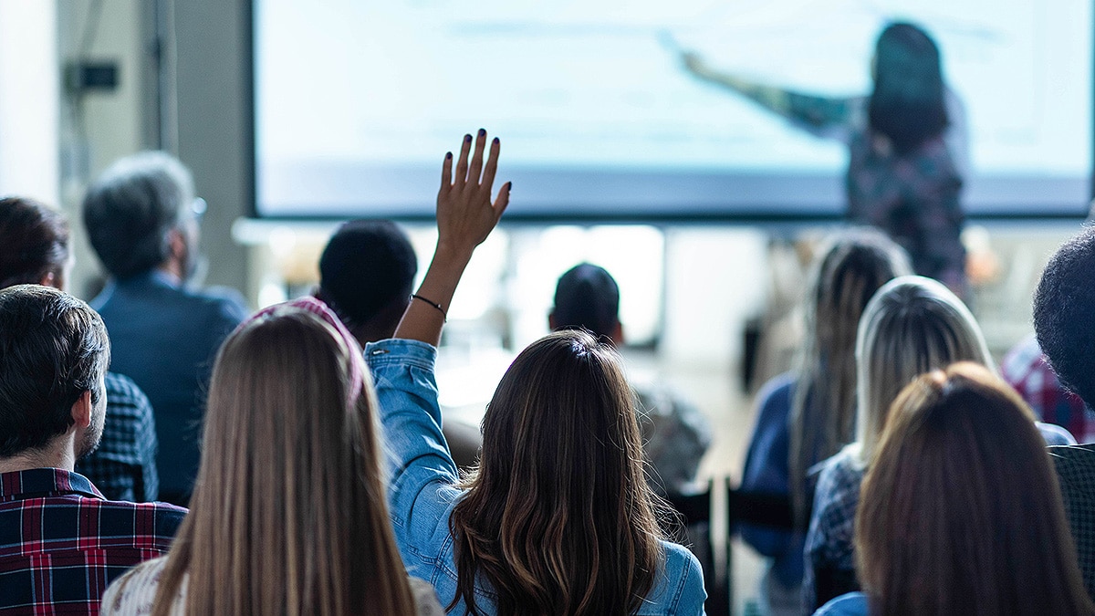 Woman at a large screen at front of room with one woman in crowd raising her hand.