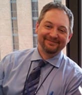 A picture of Gregg Brandush smiling while wearing a blue button up shirt and striped tie.