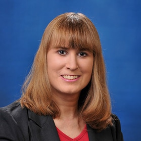 April Veoukas' headshot. She has shoulder length brown hair and is wearing a grey suit jacket with a red shirt.