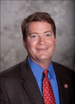 Dr. Watkins' headshot. He has short brown hair and is wearing a brown suit, blue shirt, and red tie.