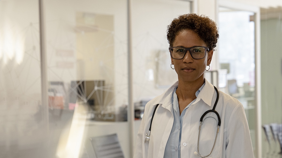 A female doctor in a white coat stands while wearing a stethoscope around her neck.