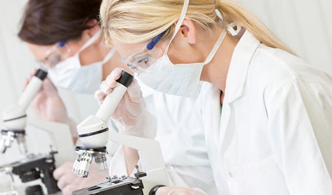 Two lab technicians wearing white lab coats, masks, and protective glasses looking through microscopes.