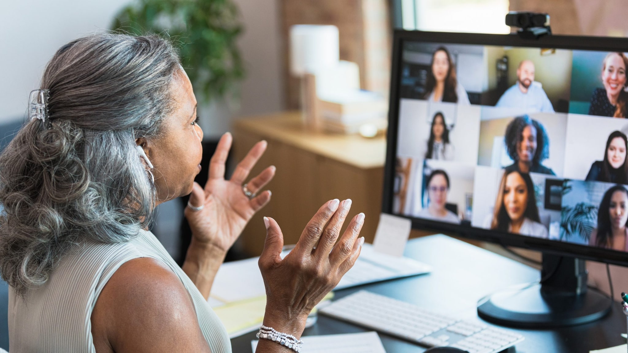 A woman sits in front of a computer and gives a virtual presentation to peers viewing on the screen.