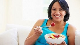 Mujer con sobrepeso sentada en el sofá comiendo un plato de frutas frescas.