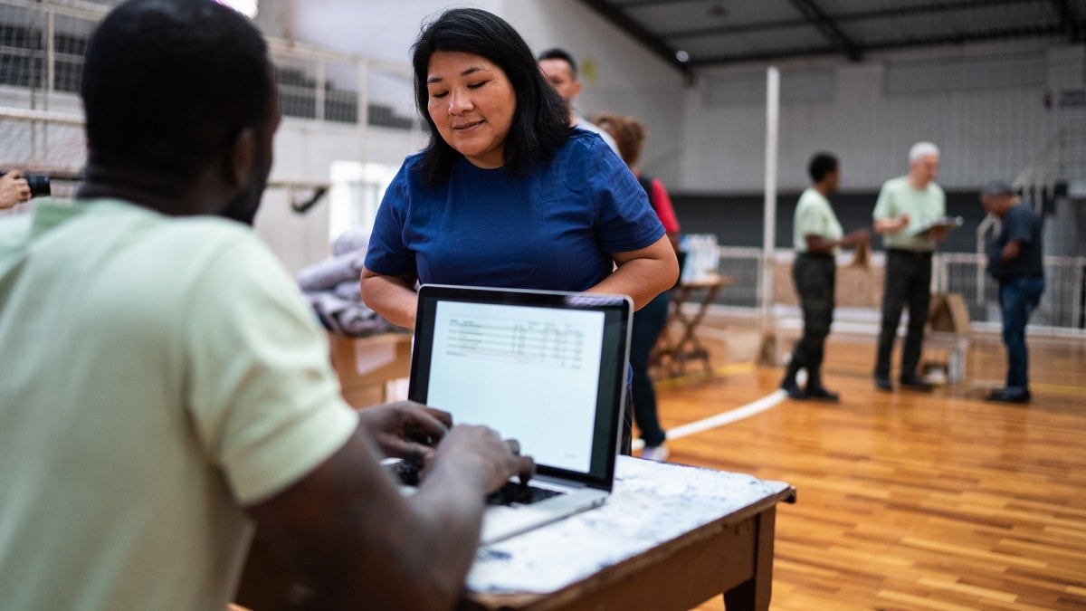 Volunteer talking with a woman while working on a laptop at an emergency shelter with others in the background.