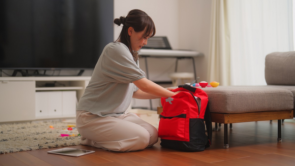 A woman kneels on the floor while packing a red and black backpack, with toys scattered around and a tablet on the floor beside her.