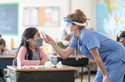 Young girl in classroom wearing mask getting temperature checked by health professional wearing PPE