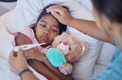 Sick young girl in bed with teddy bear getting temperature checked by caregiver.