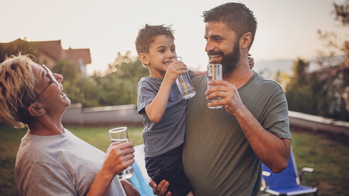 Family of two adults and one child laughing and drinking water out of glasses in the backyard