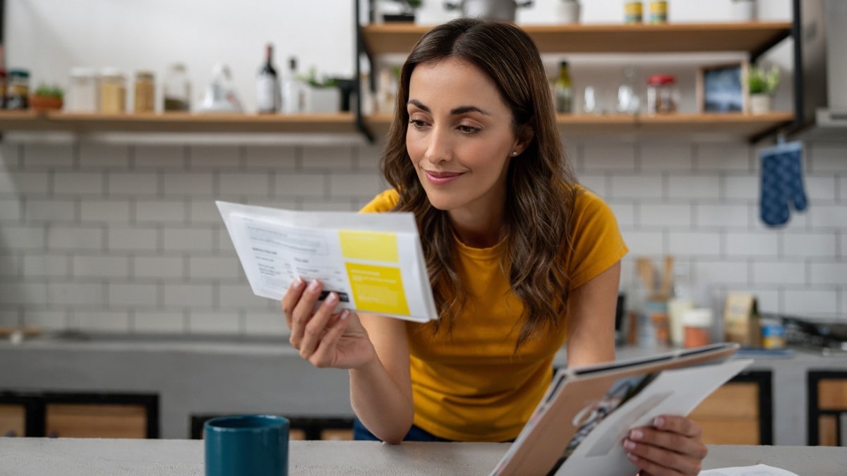 Woman in her kitchen holding mail and looking at a document.