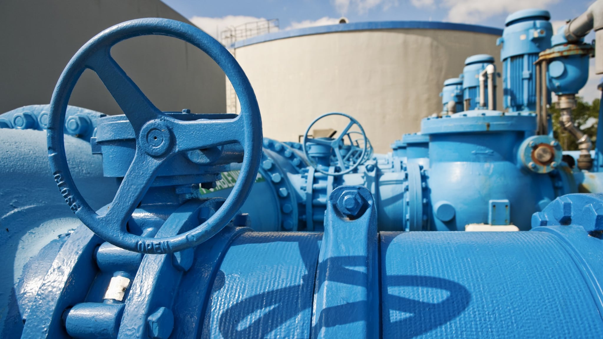 Pipes and pumps used to move water at a treatment plant in the foreground, and large water tanks in the background