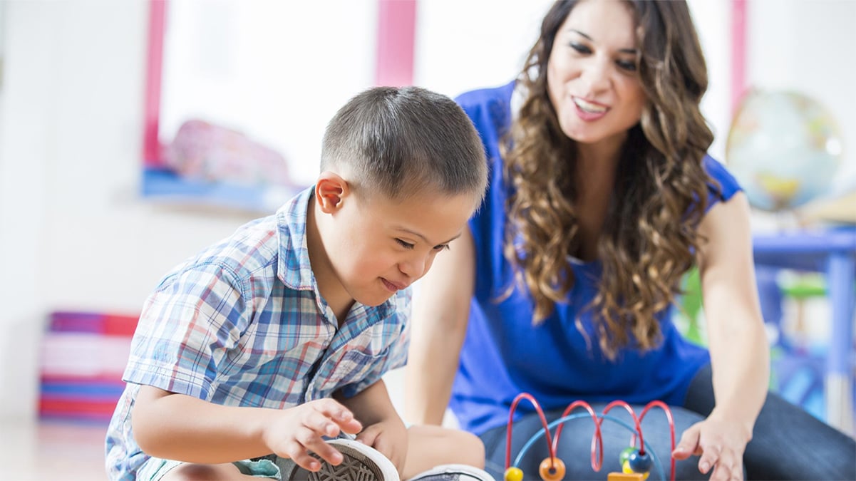 An early care and education provider sits with boy on floor who is playing with toys.