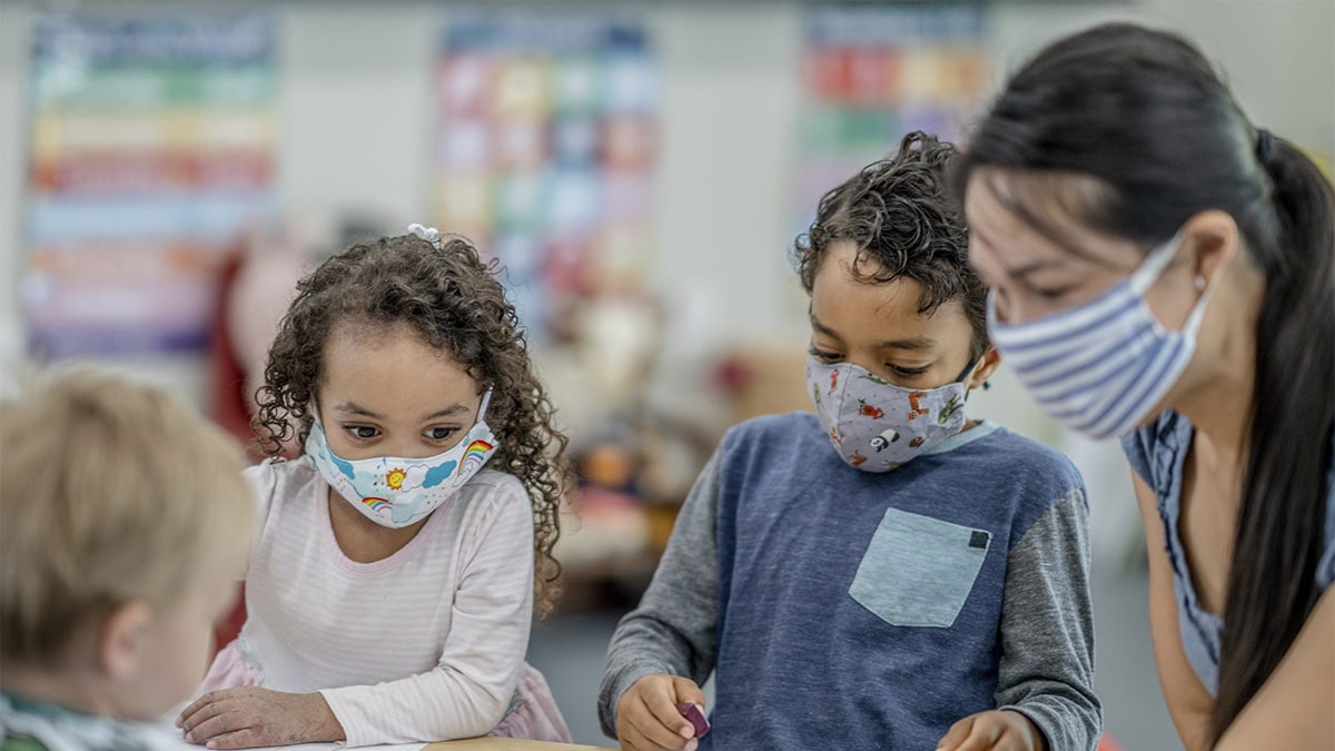 Teacher and young students wearing masks while they sit at a table together.