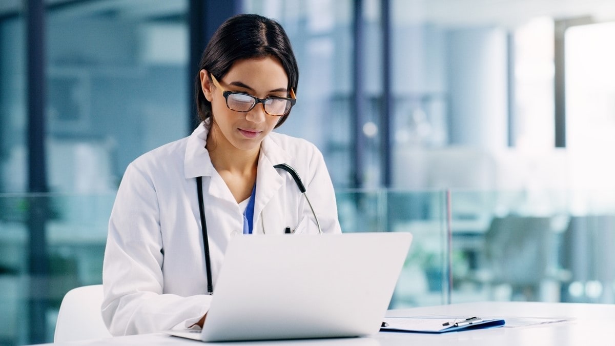 A healthcare provider wearing glasses sits at a desk with an open notebook computer.