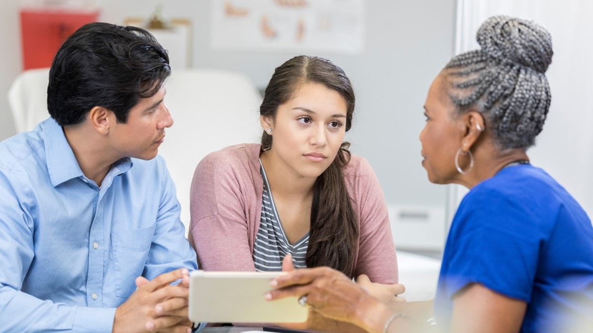 A teenage girl sits at a table next to a male, with a female healthcare provider on the other side.