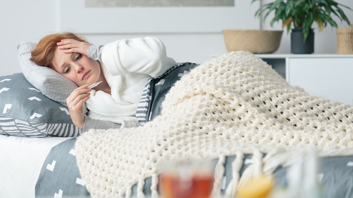 A sick woman lies in bed, looking at a thermometer and holding her hand on her head.