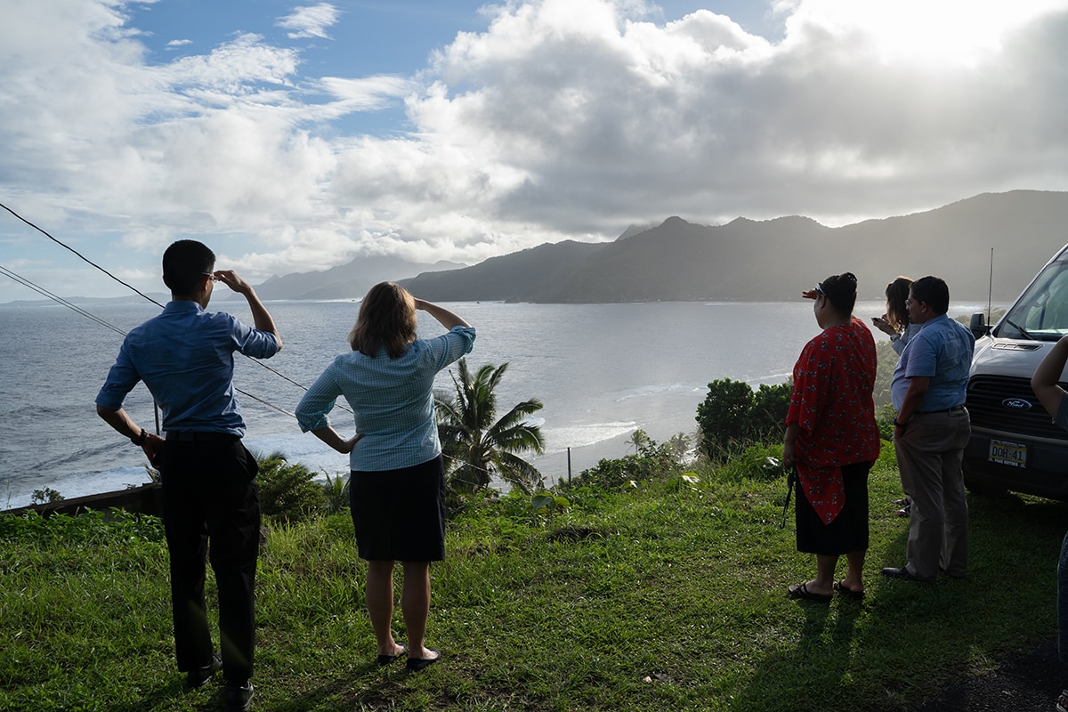 5 people stand with their backs to the camera, facing an ocean overlook with palm trees. Two of the people salute toward the view.