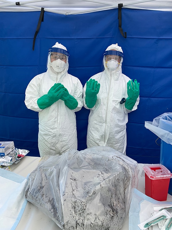 Two people in white biohazard suits stand behind a table. On the table is a multi-gallon plastic bag with unknown material inside.