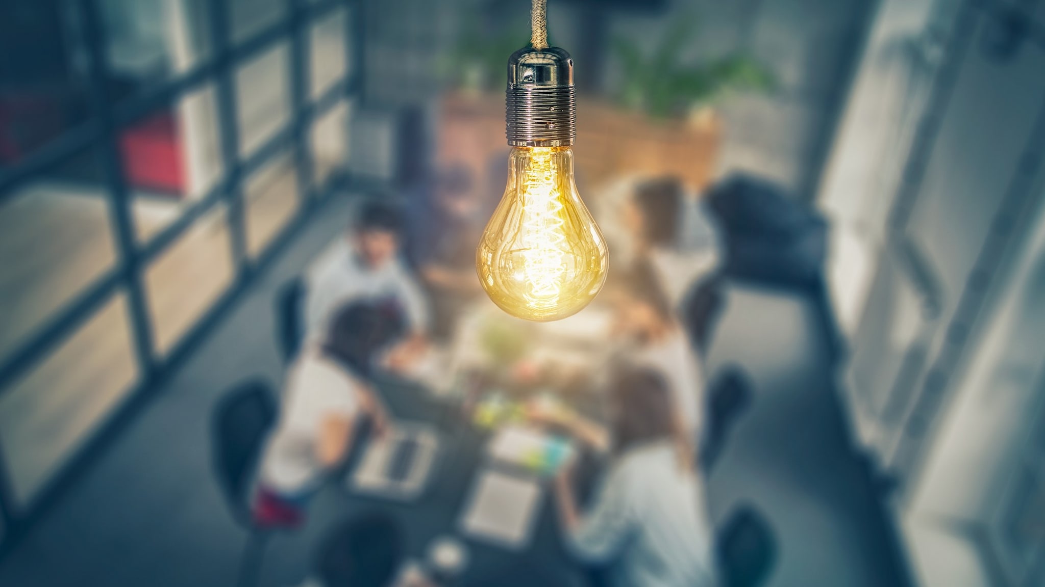 Photograph of a light bulb hanging from a ceiling with people meeting at a table in the background.