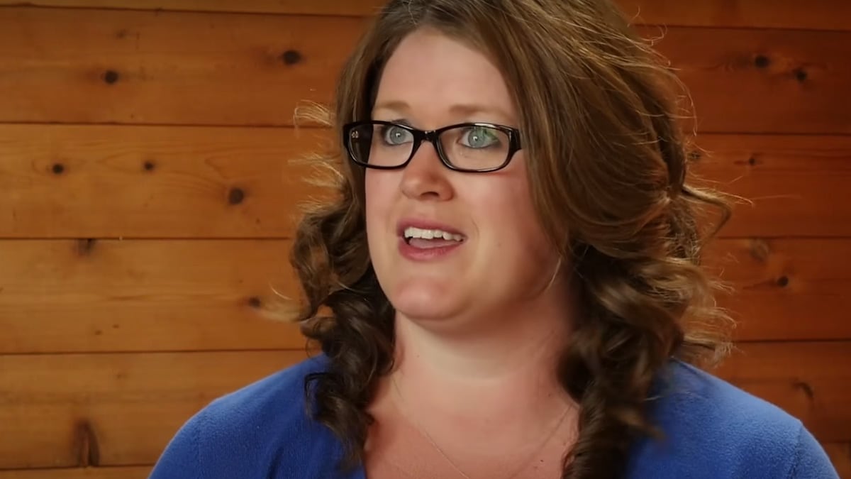 Woman wearing glasses and blue shirt with long brown hair speaking with wood-walled background.