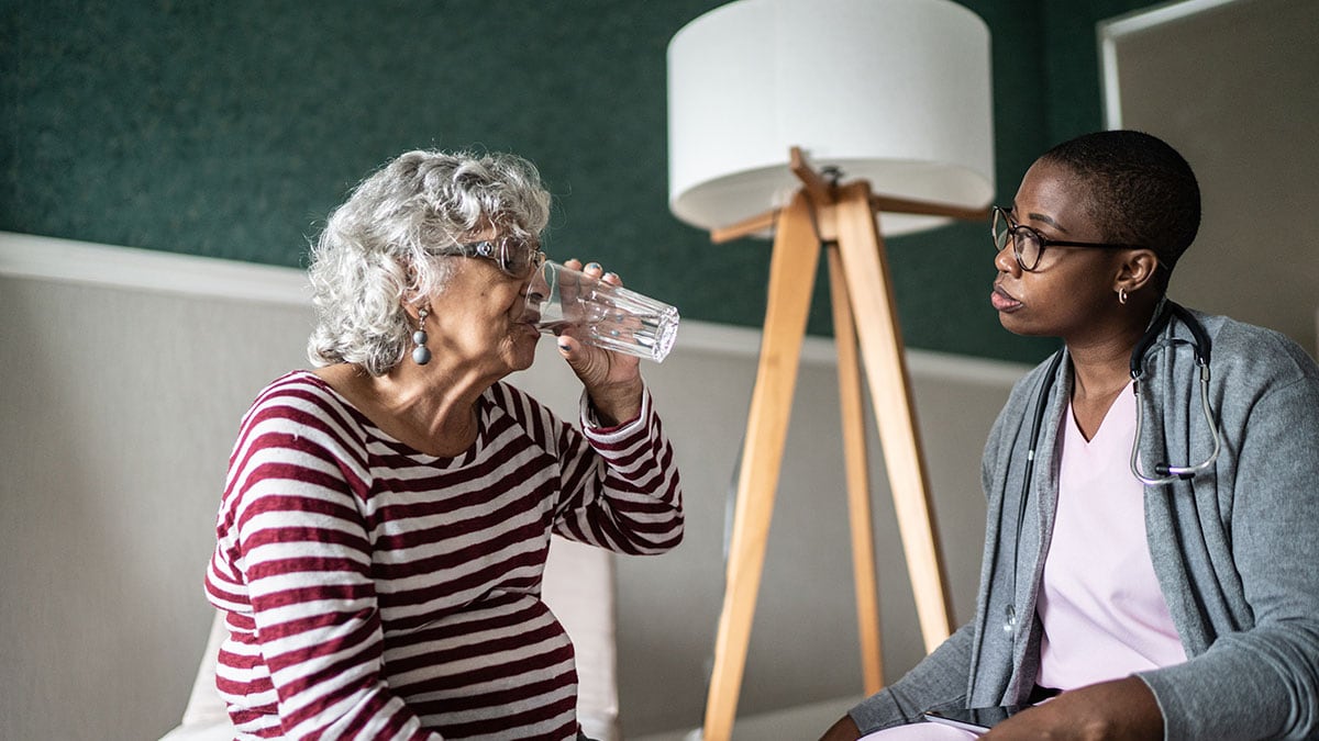 Older woman sitting inside, drinking from a glass of water while her caretaker sits beside her.