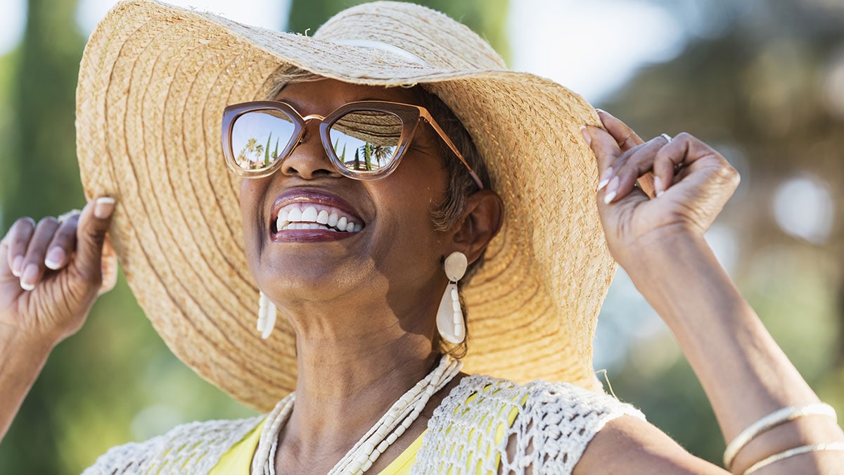 Older woman smiling as she is outside wearing sunglasses and a wide brimmed hat.