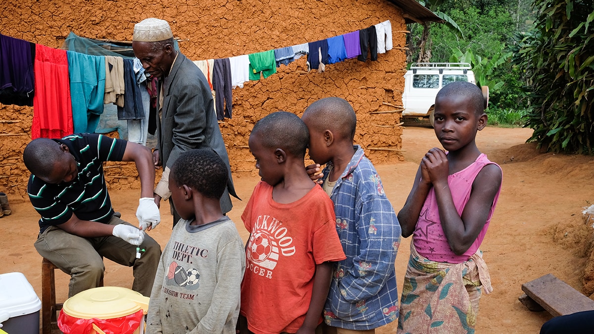 Community health worker takes blood sample via fingerstick from man while children watch on.