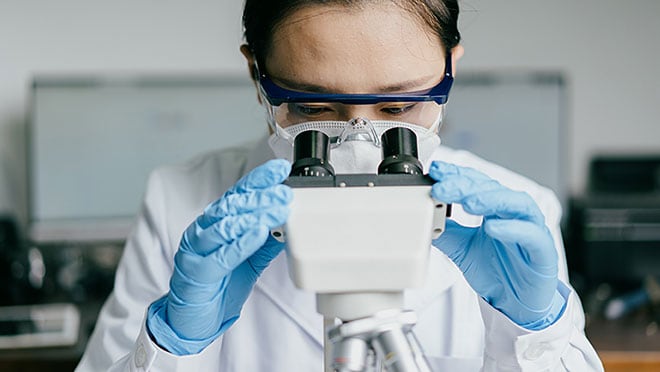 A person wearing a lab coat, safety goggles, a face mask, and blue latex gloves.