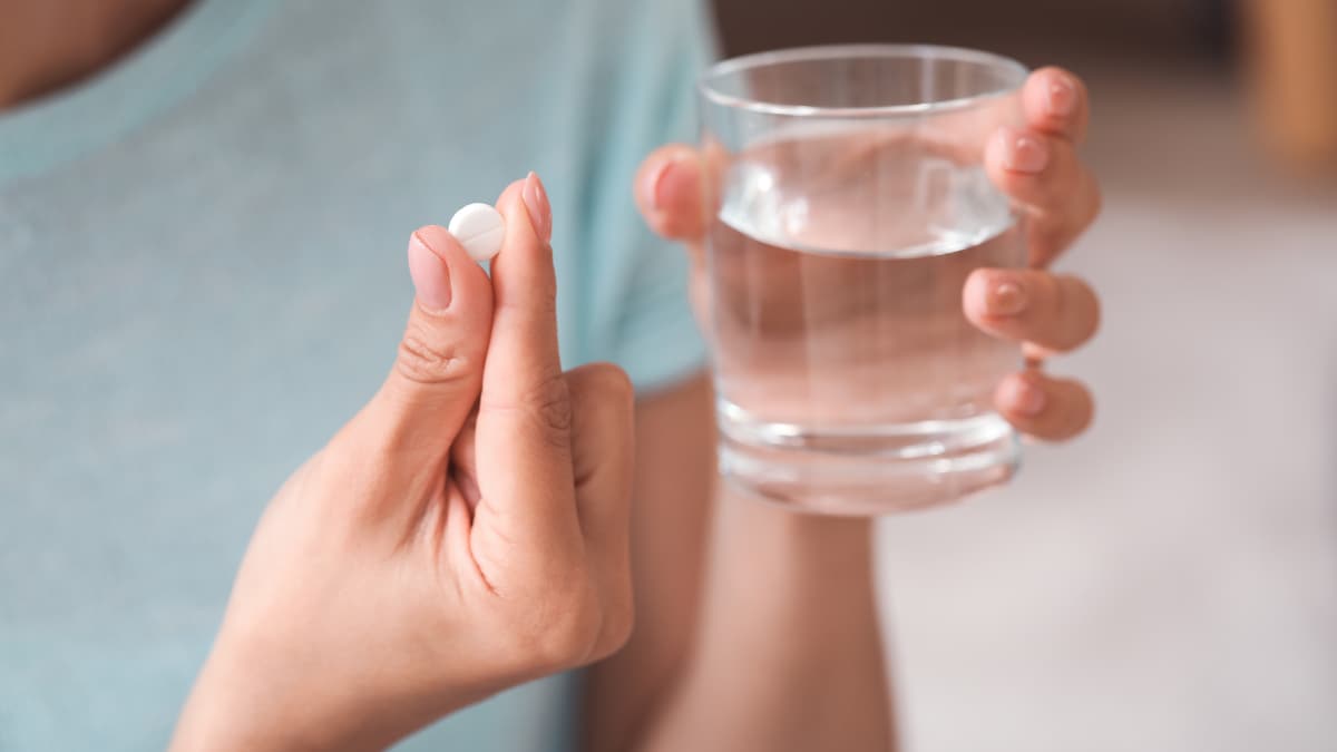 Vista de cerca de una tableta de vitamina blanca y pequeña y un vaso de agua en la mano de una persona.