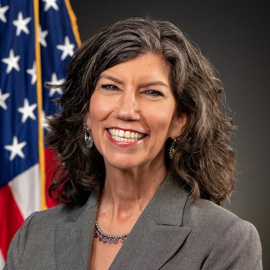 Woman with dark gray hair against a gray background with the U.S. flag.