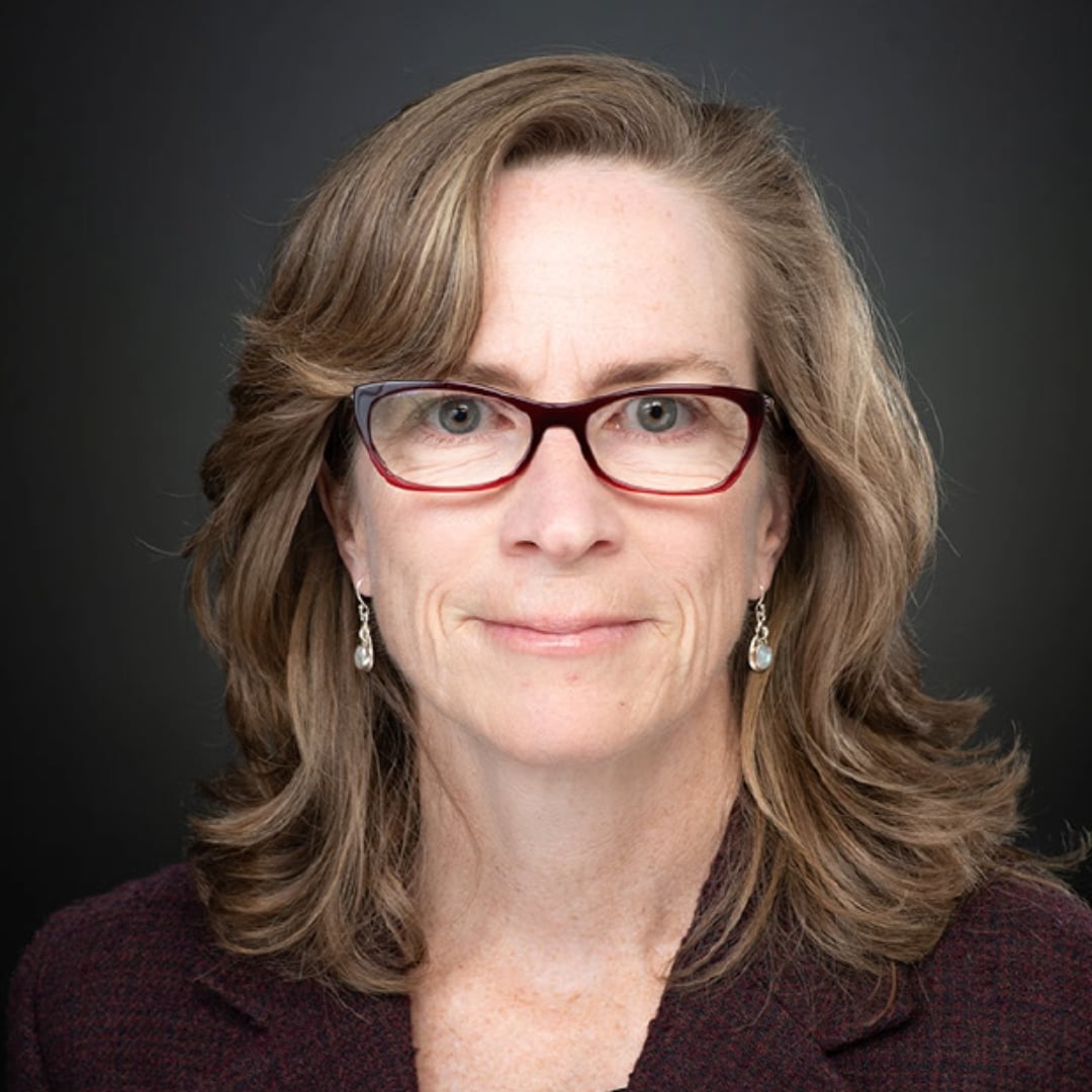 Woman with brown hair and glasses in a suit smiling.