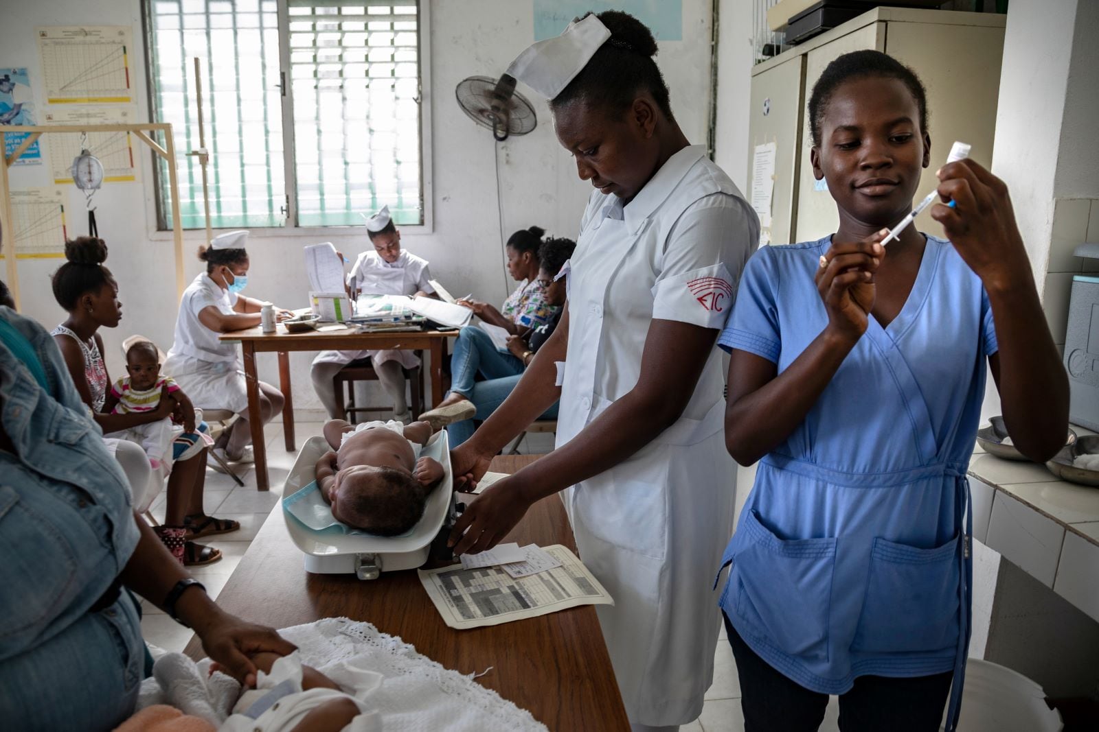 Nurse weights infant on a scale, while another nurse prepares a vaccine shot.