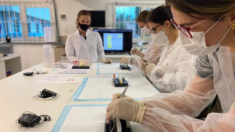 Lab technicians in masks and gloves analyze samples at a lab bench. Photo by Filipe Perini/CDC.