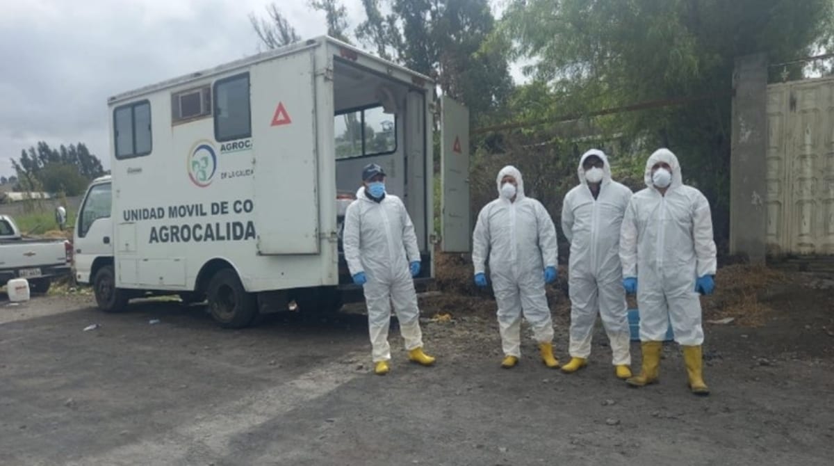 Workers are wearing white full-body protective suits, gloves, masks, and yellow boots. They are standing beside a white mobile unit labeled "Unidad Móvil de Control Agrocalidad."