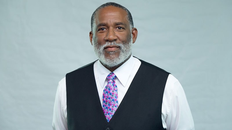 A man with a graying beard, wearing a white shirt, black vest, and a colorful floral tie, with a plain background.