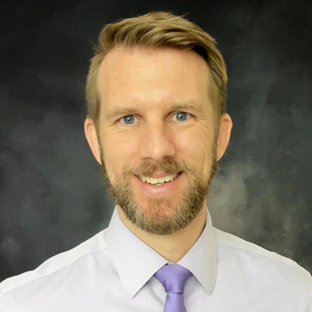 A smiling man with short hair, a beard, and a lavender tie against a dark background.