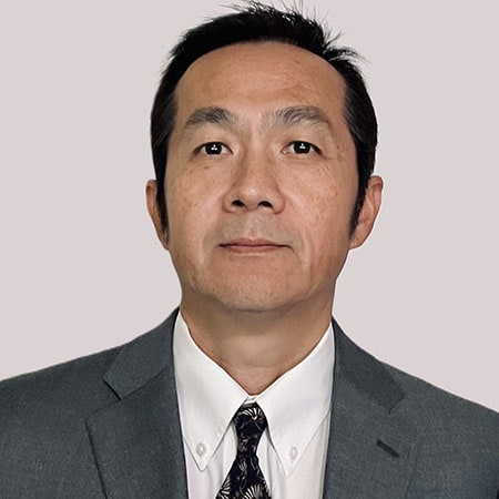 A man in formal attire, wearing a grey suit, white shirt, and dark tie, with a neutral expression.