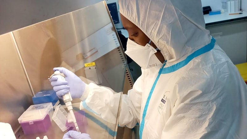 Woman in white lab coat that covers her head with face mask and purple gloves tests mpox.