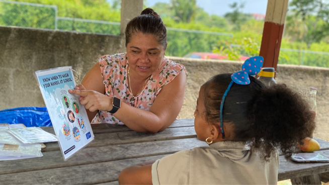 An adult sits across from a young student and points to a poster that reads "Know When to Wash Your Hands at School.