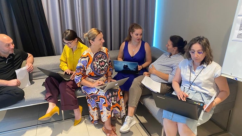 Six individuals sit on a couch talking to one another during a SIMEX exercise. Two people hold papers and four people have laptops.