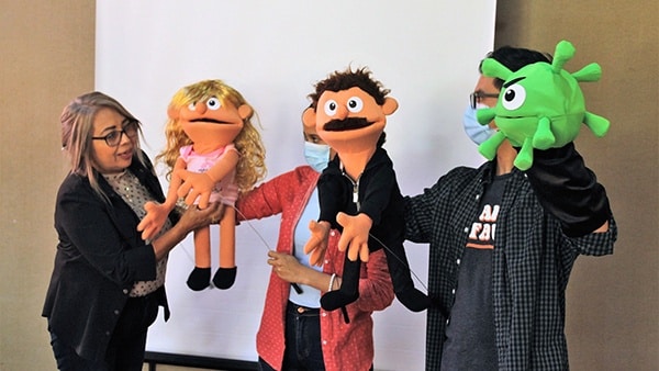 Three individuals hold hand puppets to demonstrate why pregnant women should get the COVID-19 vaccine. The three puppets resemble a coronavirus with eyes and a mouth, a woman, and a man.