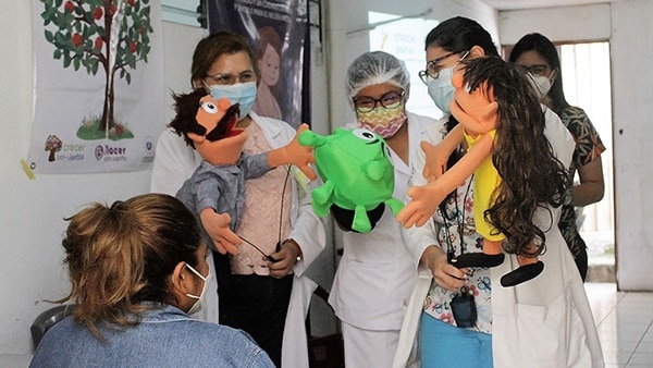 Three health workers with face masks each hold hand puppets, each resembling a child, a man, and a coronavirus with eyes and a mouth, while talking to a pregnant woman.