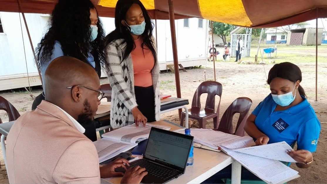 Four people stand and sit around a table under an outdoor tent.